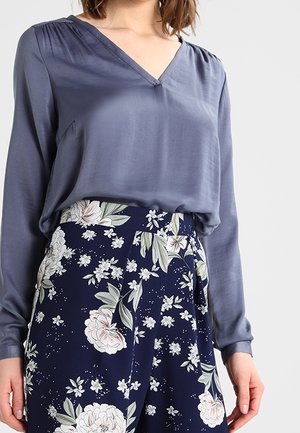 Vrouw draagt een blauwe satijnen blouse met lange mouwen, ingestopt in marineblauwe broek met een wit-groen bloemenpatroon.
