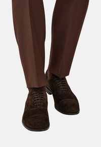 Scarpe oxford in suede marrone scuro indossate con pantaloni eleganti marroni e calzini a fantasia, mostrate dalla metà polpaccio in giù su uno sfondo chiaro.