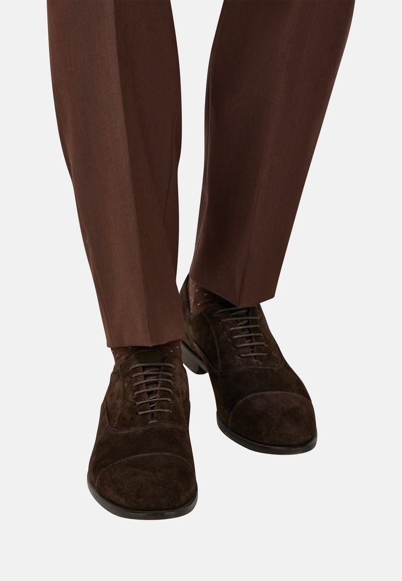 Scarpe oxford in suede marrone scuro indossate con pantaloni eleganti marroni e calzini a fantasia, mostrate dalla metà polpaccio in giù su uno sfondo chiaro.