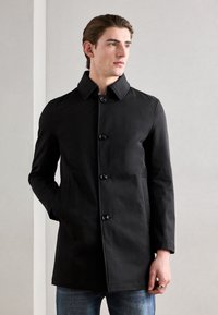 Cappotto nero con colletto alto, chiusura frontale a sei bottoni e tasche laterali. Tessuto liscio, vestibilità su misura, indossato sopra jeans blu.