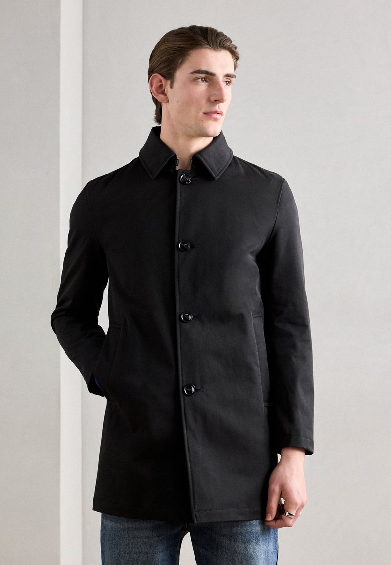 Black coat with a high collar, six-button front closure, and side pockets. Smooth fabric texture, tailored fit, worn over blue jeans.