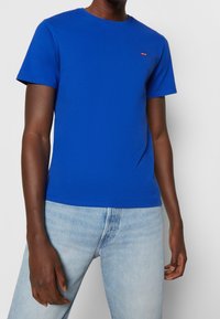 Blue cotton T-shirt with short sleeves and crew neck, featuring a small red logo on the chest. Paired with light-wash denim jeans.
