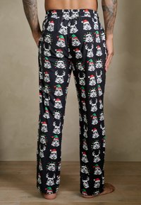 Fekete pizsamanadrág, amelyen ismétlődő minta található: rajzfilmszerű rohamosztagos arcok agancsokkal és zöld-piros karácsonyi sapkákkal.