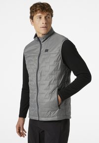 Leichte, graue Daunenweste mit einem Frontreißverschluss, featuring einem gesteppte Design, hohem Kragen und seitlichen Taschen, getragen über einem schwarzen Langarmshirt.