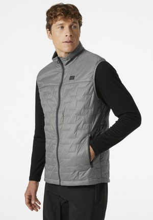 Chaleco ligero y gris tipo puffy con cremallera frontal, que presenta un diseño acolchado, cuello alto y bolsillos laterales, llevado sobre una camiseta negra de manga larga.