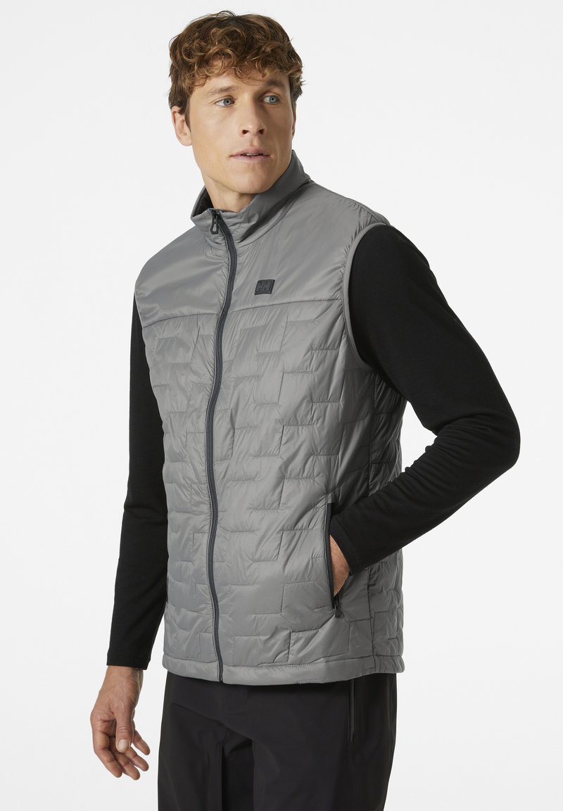 Leichte, graue Daunenweste mit einem Frontreißverschluss, featuring einem gesteppte Design, hohem Kragen und seitlichen Taschen, getragen über einem schwarzen Langarmshirt.