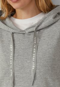 Grijze hoodie met een zachte textuur, voorzien van een trekkoord met de tekst "liefde staat aan jouw zijde" en een witte binnenkant van de kraag.