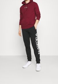 Sudadera burdeos con cordones y bolsillo delantero, combinada con pantalones de chándal negros que presentan el logo blanco de "Nautica". Zapatillas blancas completan el atuendo.