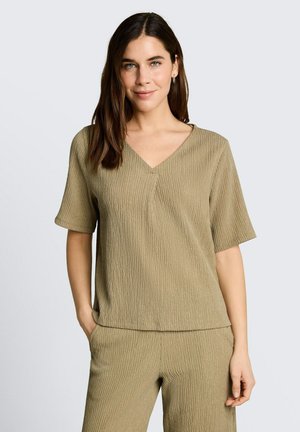 Kobieta o brązowych włosach, ubrana w teksturowany oliwkowozielony top z krótkim rękawem i dekoltem w serek oraz pasujące spodnie, stoi z jedną ręką w kieszeni.