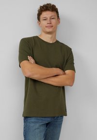 Oliwkowa, krótki rękaw t-shirt z fakturowanej dzianiny. Okrągły dekolt i luźny krój. Noszony z klasycznymi dżinsami.