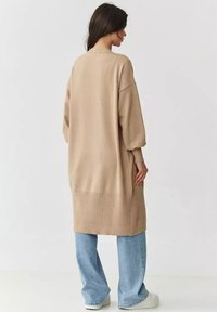 Beżowy, oversize sweter z dzianiny z prążkowanym dołem i rękawami, zestawiony z jasnoniebieskimi spodniami o szerokich nogawkach i białymi trampkami.