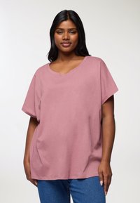 Jasnoróżowa koszulka z krótkim rękawem, o szerokim dekolcie i luźnym kroju, uszyta z miękkiego materiału. W połączeniu z niebieskimi dżinsami.