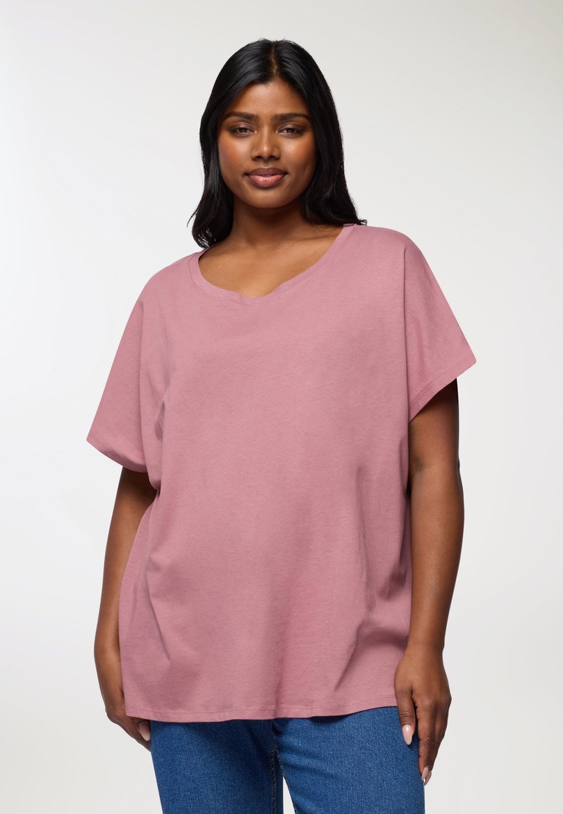 Jasnoróżowa koszulka z krótkim rękawem, o szerokim dekolcie i luźnym kroju, uszyta z miękkiego materiału. W połączeniu z niebieskimi dżinsami.