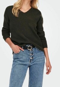 Pull en maille vert foncé avec un décolleté en V associé à un jean en denim bleu taille haute avec une fermeture à cinq boutons et une ceinture noire avec une accent en or.