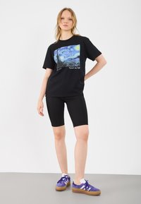 Camiseta preta com uma impressão gráfica azul e amarela retratando um céu estrelado. Combinada com calções de ciclismo pretos e ténis roxos.