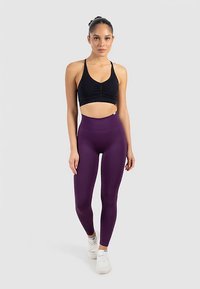 Reggiseno sportivo nero con centro arricciato, abbinato a leggings viola a vita alta e scarpe sportive bianche, su uno sfondo neutro.