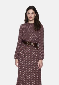 Langärmeliges, kastanienbraunes Kleid mit einem geometrischen Muster in Creme und Rot. Es verfügt über einen hohen Ausschnitt und einen braunen Gürtel mit einer runden Schnalle.