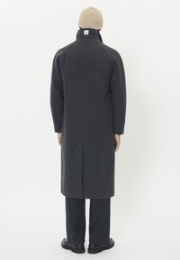 Lungo cappotto di lana grigia con colletto alto e spacco posteriore, indossato sopra pantaloni neri e scarpe nere. Accompagnato da un berretto a maglia beige.