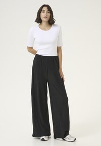 Biely priliehavý top s krátkym rukávom v kombinácii s čiernymi zvrásnenými širokými nohavicami so elastickým pásom. Model má obuté biele športové topánky.