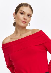 Roter off-shoulder Pullover mit geripptem Halsauschnitt und glatter Textur. Hat lange Ärmel, einen taillierten Schnitt und weichen Stoff.