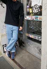 Černá košile s dlouhým rukávem, světle modré volné džíny a tmavě modré tenisky s oranžovými detaily. V pozadí je skleněné dveře pokryté graffity.