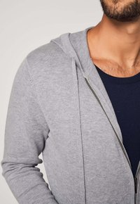 Felpa grigia con cerniera a zip e una lavorazione a trama, dotata di cappuccio e cerniera argento. Indossata sopra una maglia a girocollo blu scuro.