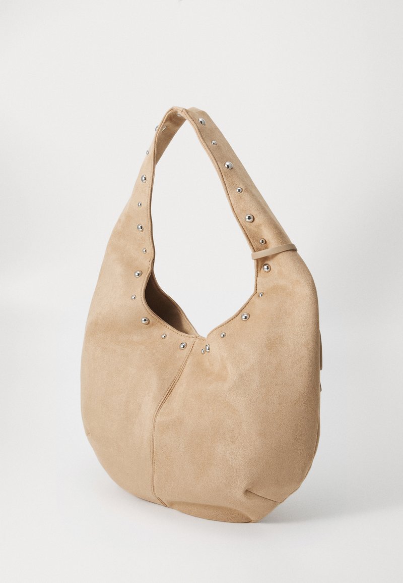 Bolso hobo de ante beige con forma redondeada, que cuenta con un asa superior y tachuelas plateadas a lo largo de la abertura para un detalle adicional.