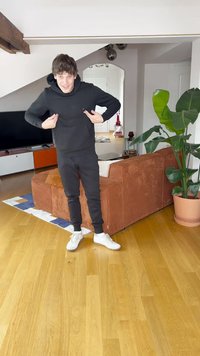 Czarna bluza z kapturem i spodnie dresowe, białe trampki, wnętrze z brązową kanapą z sztruksu, drewnianą podłogą oraz dużą zieloną rośliną doniczkową.