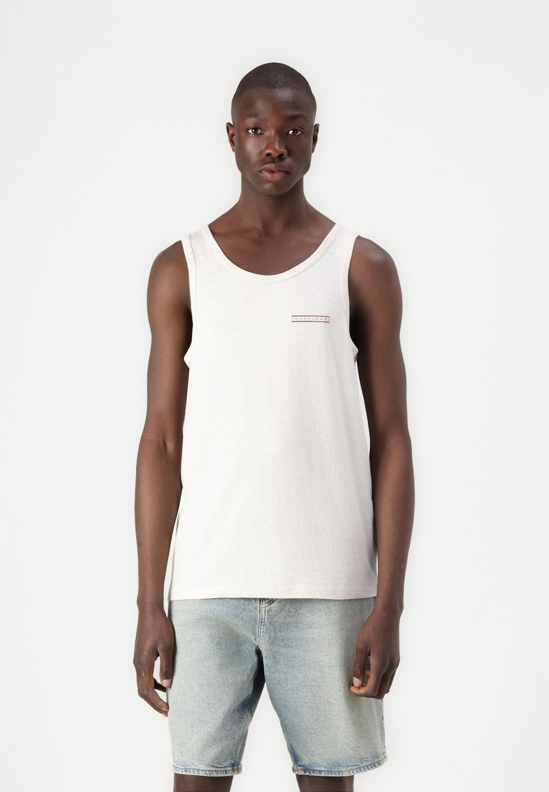 Canotta grigia chiara con scollatura rotonda, caratterizzata da un piccolo logo grafico orizzontale rosso e nero sul petto. Indossata con pantaloni corti in denim blu.