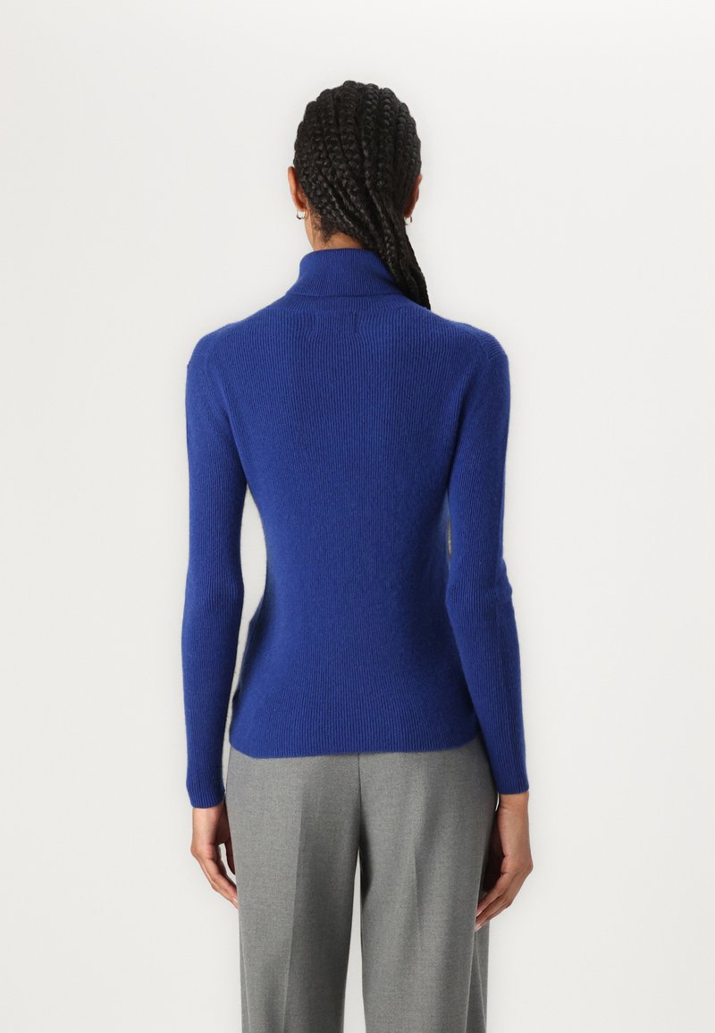 Suéter de cuello alto azul cobalto con textura acanalada, silueta ajustada y mangas largas, visto desde la parte trasera. Combinado con pantalones grises.
