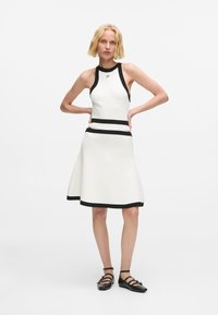 Fehér ujjatlan ruha fekete díszítéssel és bővülő szegéllyel. Magas nyakkivágással és vízszintes csíkokkal a deréknál és a szegélyén.