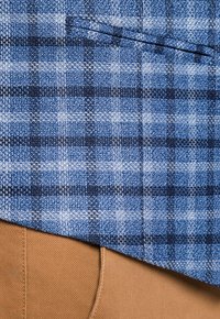 Niebiesko-czarna tkanina w kratę z tkaną teksturą leży na jasnobeżowej tkaninie. Posiada kieszeń na piersi w górnej części.