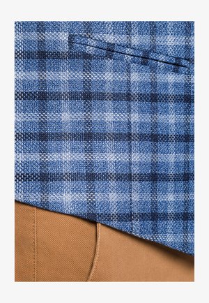 Niebiesko-czarna tkanina w kratę z tkaną teksturą leży na jasnobeżowej tkaninie. Posiada kieszeń na piersi w górnej części.