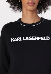Melna sporta krekls ar baltu "KARL LAGERFELD" uzrakstu, noapaļota apkakle ar kontrastējošu baltu apdari, gluda tekstūra un brīvs piegriezums.