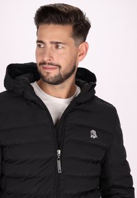 Melna pūka jaka ar rāvējslēdzēju, kapuci un sānu kabatām. Izceļas ar diskreto logo uz krūtīm un quilted tekstūru.