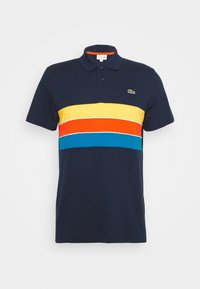 Tmavě modré polotričko s žebrovaným límcem. Na hrudi má vodorovné pruhy ve žluté, oranžové a modré barvě. Na levé straně je logo Lacoste.