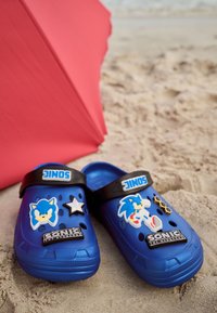 Sabots bleus pour enfants avec des motifs Sonic the Hedgehog sur le sable à côté d'un parasol rouge de plage.