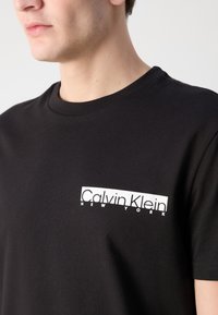 Černé bavlněné tričko s bílým obdélníkovým logem s textem "Calvin Klein NEW YORK". Klasický crew neck a krátké rukávy.