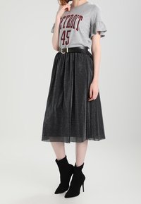 Kvinna iklädd en grå t-shirt med rynkade ärmar "Detroit 45" instoppad i en glittrig svart midi-kjol med svart bälte och svarta högklackade ankelstövlar.