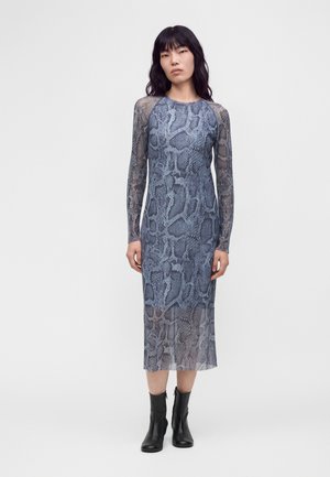 Femme aux longs cheveux noirs portant une robe midi à manches longues imprimée serpent bleu et gris et des bottines noires, debout sur un fond uni.