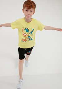 Garçon roux en équilibre sur une jambe, portant un t-shirt jaune à motif dinosaure, un short noir avec un motif bleu et des baskets blanches sur fond blanc.