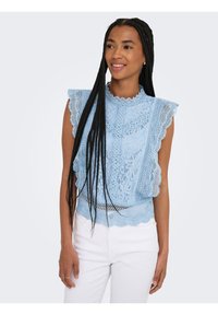 Femme aux longues tresses portant un haut en dentelle bleu sans manches et un pantalon blanc, souriante et regardant légèrement sur le côté.