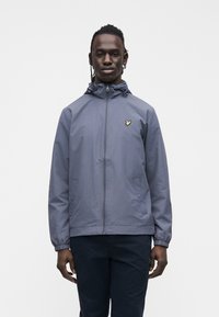 Man wearing a gray zip-up hooded jacket with a yellow bird logo on the chest and dark pants, standing against a plain background.