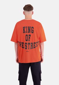 Orange Baumwoll-T-Shirt mit schwarzem Text "KING OF THE STREET" auf dem Rücken. Kurze Ärmel, lockere Passform und runder Ausschnitt.