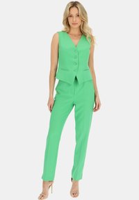 Zielona dopasowana kamizelka z dekoltem w kształcie litery V, pięcioma guzikami i dwiema przednimi kieszeniami, w połączeniu z pasującymi wąskimi spodniami, oba z teksturowanej tkaniny.