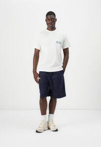 White cotton t-shirt with a small blue graphic on the chest, paired with navy blue shorts and beige sneakers with white accents.
