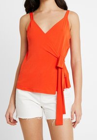 Femme portant un haut sans manches enroulé orange vif avec un nœud sur le côté et un short en denim blanc effiloché devant un fond blanc uni.