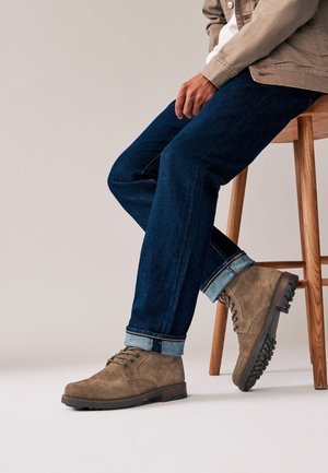 Braune Wildleder-Stiefeletten mit Schnürdesign und robuster Sohle, kombiniert mit hochgekrempelten dunkelblauen Jeans. Holzstuhl im Hintergrund.