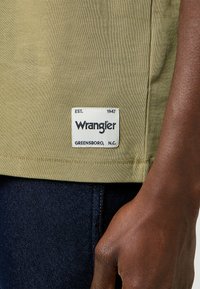 T-shirt in cotone verde oliva con un'etichetta rettangolare bianca con la scritta "Wrangler Est. 1947 Greensboro, N.C." cucita all'orlo, abbinato a jeans scuri.
