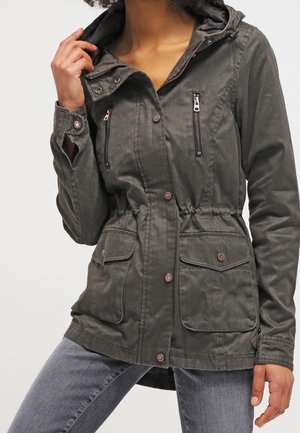 Woman wearing a dark olive green hooded jacket with front zipper and button pockets, paired with faded gray jeans.
